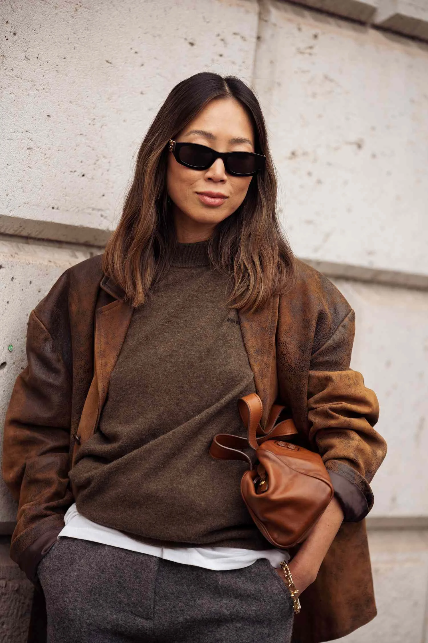 Aimee Song wears brown jumper, leather jacket, grey pants, brown bag outside MiuMiu show during the Womenswear Spring Summer 2026