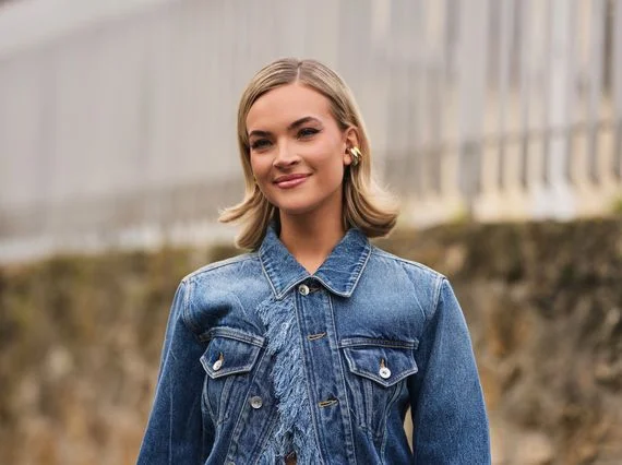 A woman wearing a denim jacket standing outdoors with a neutral expression